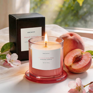 Peach Oolong soy candle lit beside its packaging box, with a fresh peach and cherry blossom sprigs
